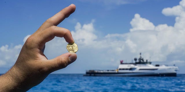 Een ontdekkingsreiziger houdt een gouden munt vast die gevonden is op de Bahama's, waar de verkenningsboot van de Allen al van ver te zien is.
