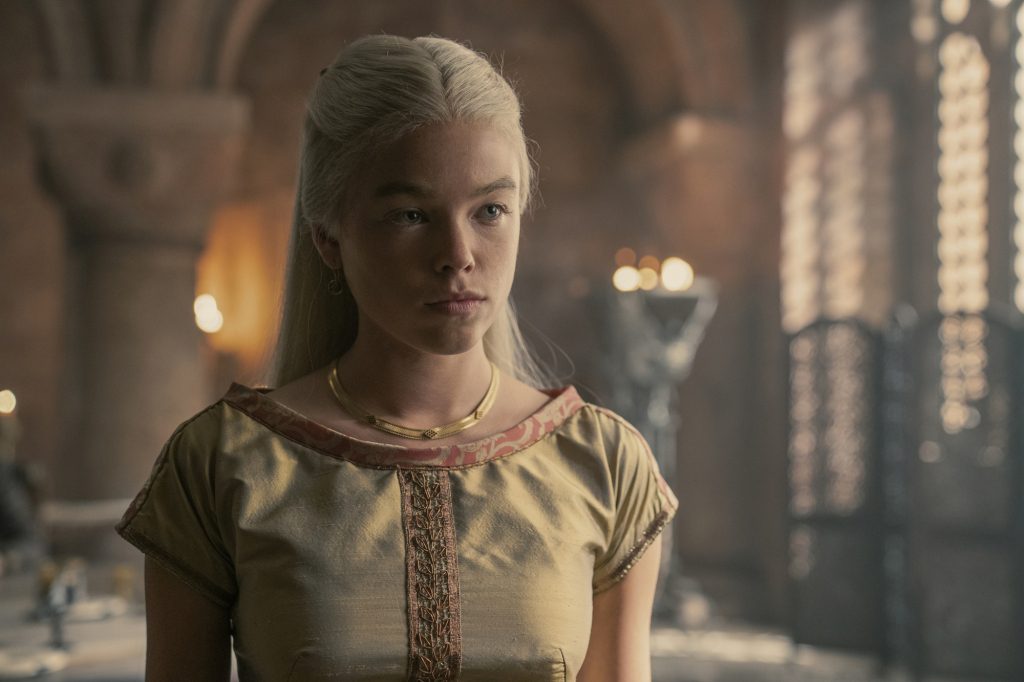 Millie Alcock als prinses Rhaenyra Targaryen in "Drakenhuis" Staande in een kamer, gekleed in een jurk. 
