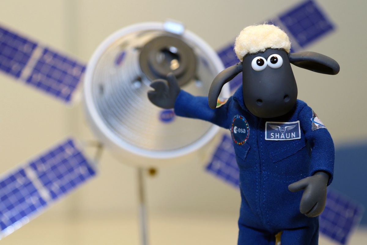 A plush cartoon sheep holds a hoof against a small model of an Orion capsule.