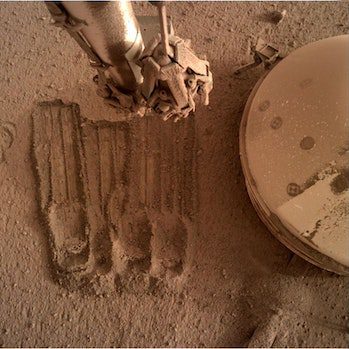 Op een andere recente foto gebruikt InSight zijn robotarmen om een ​​deel van de omtrek van de regoliet te verwijderen...