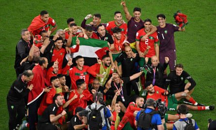 Een groep spelers in hun rode shirts verzamelt zich voor een foto op het veld, gebarend ter ere van de viering.  Verschillende spelers hijsen de vlag van Palestina in het midden van de groep